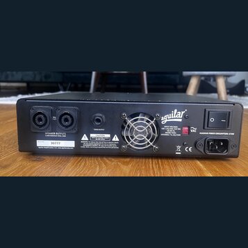 Zadní strana černého basového zesilovače značky aguilar. Jsou zde dva výstupy SPEAKER OUTPUTS s nápisem 4 OHM MINIMUM TOTAL LOAD, výstup TUNER OUTPUT…