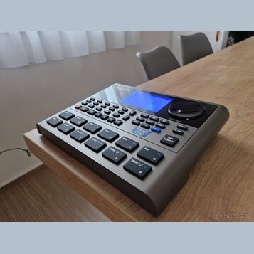 Šedý elektronický bicí modul s modrým displejem a velkým ovládacím knoflíkem. Na horní části je viditelné logo Roland. Zařízení obsahuje množství gum…