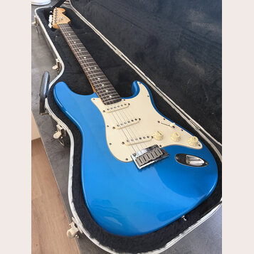 Modrá elektrická kytara typu Stratocaster s krémovým pickguardem a hnedým hmatníkem, umístěná v otevřeném tvrdém kufru. Na hlavě krku je viditelné lo…