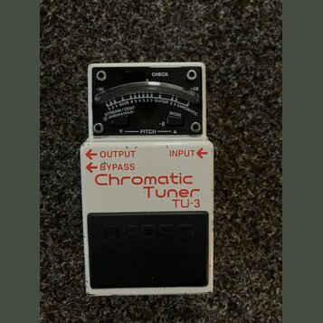 Bílý kytarový ladič s černým spínacím pedálem. Na spodní části je červený nápis Chromatic Tuner TU-3 a šipky OUTPUT, BYPASS a INPUT. Horní část obsah…