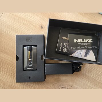 Na fotografii je malý kytarový zesilovač v originálním černém balení. Na krabici i na přístroji je bílý nápis NUX a označení modelu MP-3 MIGHTY PLUG…
