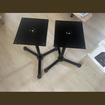 Dva černé stojany s hranatými horními deskami a trojnožkami. Na deskách jsou uprostřed kulaté otvory. V pravé části snímku je vidět část krabice s ná…