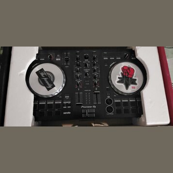 Černý DJ 컨트롤러 Pioneer Dj s logem serato v dolní části. Zařízení má dva stříbrné jog-wheels, z nichž oba jsou zakryty nálepkami; na levém je nápis The…