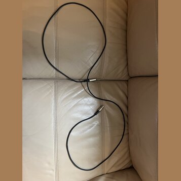 Černý instrumentální kabel s kovovými konektory. Kabel je položen na béžovém koženém povrchu. Na kabelu ani konektorech nejsou viditelné žádné značky…
