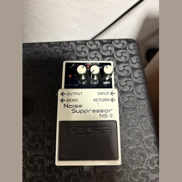 Bílý efekt pedál s černým spínacím tlačítkem. Na zařízení jsou nápisy BOSS, Noise Suppressor NS-2 a popisky ovládacích prvků REDUCTION, MODE, OUTPUT,…