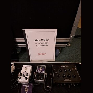 Na fotografii je otevřený transportní kufr s několika efektovými pedály a uživatelskou příručkou. Na bílém sešitě je nápis MESA/BOOGIE, M3 CARBINE. O…