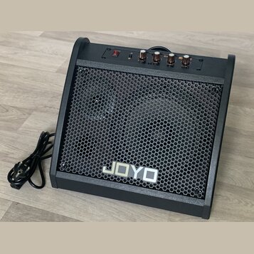 Černý kytarový kombo zesilovač s kovovou mřížkou včelíkového vzoru. Na přední straně je umístěno bílé logo JOYO. Horní panel obsahuje červený vypínač…