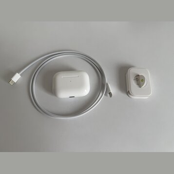 Bílé nabíjecí pouzdro k bezdrátovým sluchatkům, šedý pletený kabel s konektory USB-C a bílý síťový adaptér. Na přední straně pouzdra je vidět malá ko…