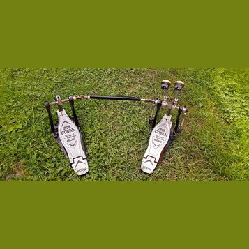 Dvojpedál na buben v černé a stříbrné barvě, umístěný na trávě. Na obou šlapkách jsou nápisy IRON COBRA, The Legend in Innovation a 600, v dolní část…
