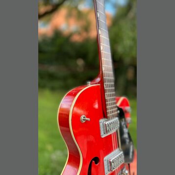 Červená polodublová elektrická kytara s bílým lemováním hran. Na těle jsou vidět dva stříbrné humbuckery a černý plecháč. Povrch laku je lesklý, bez…