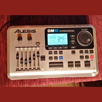 Stříbrný elektronický bicí modul značky ALESIS, model DM10. Na přední straně je displej s nápisem Fusion a Tempo 24 BPM, pět posuvníků pro ovládání h…