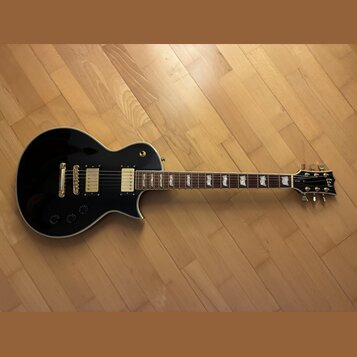 Černá elektrická kytara s tvarem singlecut a bílým lemováním. Na hlavě je viditelné logo Epiphone. Nástroj má dva humbuckery, zlatý hardware a hриф s…