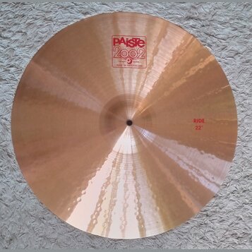 Kruhové činely z bronzu s viditelným broušením povrchu. Na horní části je červené logo Paiste 2002 s nápisy TRADE MARK a MADE IN SWITZERLAND. V pravé…