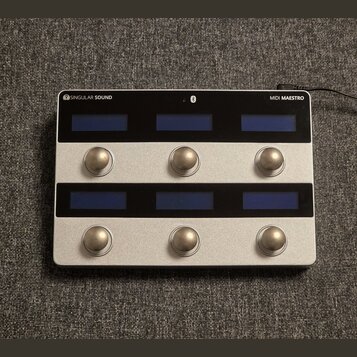 Stříbrný MIDI kontrolér s nápisy SINGULAR SOUND a MIDI MAESTRO. Zařízení má dvě řady tří modrých displejů a dvě řady tří kulatých kovových tlačítek.…