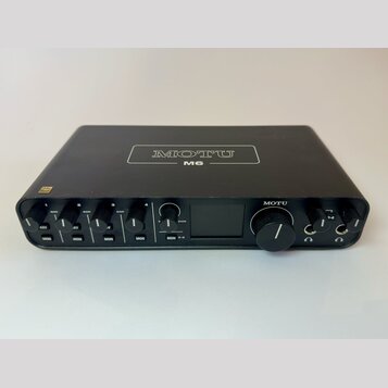 Černý audio rozhraní s nápisem MOTU M6 na horní straně. Přední panel obsahuje čtyři ovratky GAIN, tlačítka MON a jeden velký ovládací knoflík s nápis…