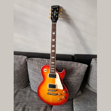 Elektrická kytara s tvarem Les Paul v červeno-žlutém sunburst provedení. Na hlavě hředu je bílý nápis Harley Benton a v dolní části logotyp s písmene…