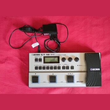 Stříbrný basový efektový procesor s černým pedálem, na kterém je nápis BOSS GT-1B BASS EFFECTS PROCESSOR. Na přístroji jsou vidět ovládací knoflíky,…