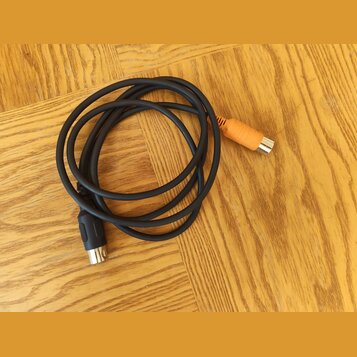 Černý audio kabel svynutý do kroužku, položený na dřevěné podlaze. Na koncích jsou dva různé konektory: jeden černý a druhý oranžový. Na kabelu nejso…