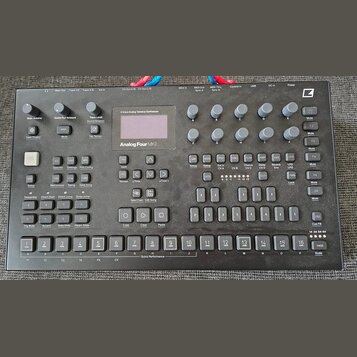 Černý analogový tabletop syntetizér s nápisem Analog Four MKII. Přístroj obsahuje displej, množství tlačítek, posuvníky a otočné knoflíky. V pravém h…