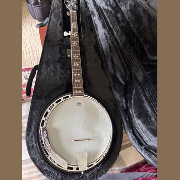 Bílá banjo v černém plišovém kufru. Na hrazence je vidět logo "Deering" a nápis "50-200 HERTZ ONLY". Hmatník je zdoben bílými vložkami ve tvaru křížk…