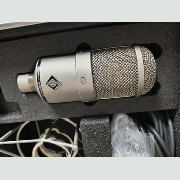Stříbrný kondenzátorový mikrofon v černé pěnové výplni kufru. Na těle je vidět logo značky a nápis NEUMANN MICROPHONE. V dolní části kufru jsou vidět…