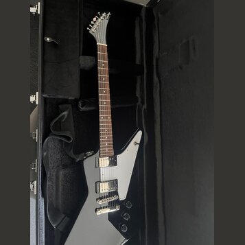 Šedá elektrická kytara s hranatým tělem a bílým pickguardem, položená v černém kufru. Nástroj má dva humbuckery a tři ovládací knoflíky. Na hlavě kyt…