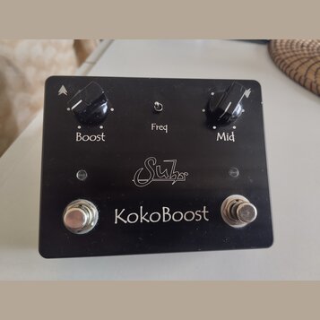 Černý kytarový efekt s nápisem KokoBoost a logem Suhr. Na horní straně jsou dva černé otočné knoflíky s popisky Boost a Mid, mezi nimiž se nachází ma…