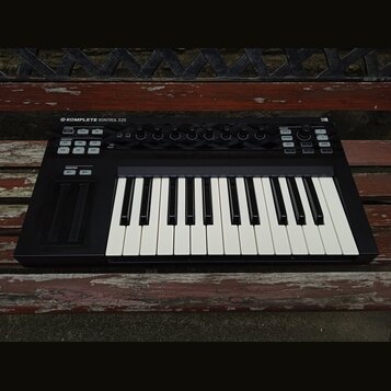 Černý MIDI kontrolér s klaviaturní klávesou. Na horním panelu je nápis KOMPLETE KONTROL S25 a v pravém horním rohu označení MK1. Zařízení obsahuje ně…