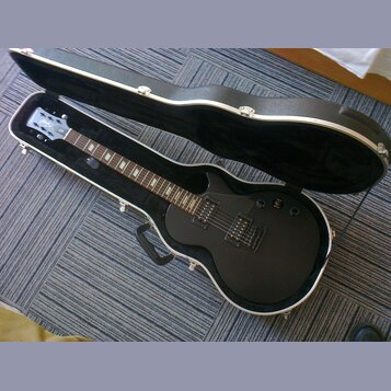 Černá elektrická kytara s tvarem singlecut v otevřeném černém tvrdém kufru. Na hlavě je viditelné logo ESP. Kytara má dva humbuckery, dvě kontrolní k…