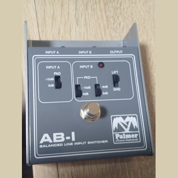 Šedý kovový přístroj s nápisem AB-I BALANCED LINE INPUT SWITCHER a logem Palmer Musical Instruments. Na horní straně jsou označení INPUT A, INPUT B a…
