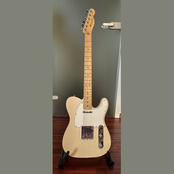 Elektrická kytara typu Telecaster v krémové barvě s bílým plechtrátem. Na hlavě hředu je viditelné logo Fender. Nástroj má javorový hmatník a dvě sní…
