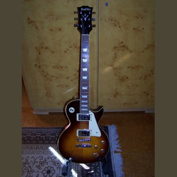 Elektrická kytara s tónem sunburst, bílým pickguardem a dvěma humbuckery. Na hlavě je nápis BaCha a pod ním logo ve tvaru stylizovaného písmena V. Hр…