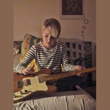 Dítě v proužkovaném tričku drží v rukou elektrickou kytaru s přírodním dřevěným povrchem těla. Kytara má bílý pickguard, dva snímače a tři ovládací k…