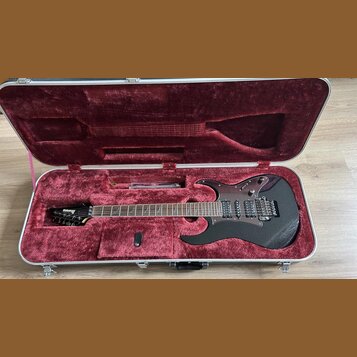 Černá elektrická kytara v hliníkovém kufru s červeným plyšovým vnitřkem. Na hlavě nástroje je bílý nápis Ibanez. Kytara má dva humbuckery, tremolo mo…