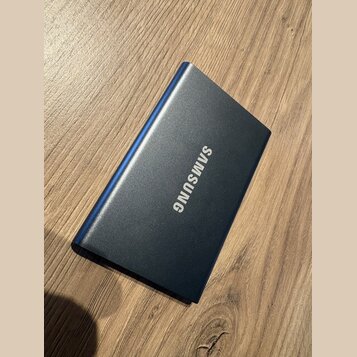 Na fotografii je modrý obdélníkový předmět s kovovým povrchem, který leží na dřevěném stole. Uprostřed je bílý nápis SAMSUNG. Povrch předmětu vypadá…