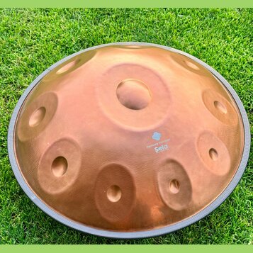 Měděný handpan s několika vyhlubenými tóny, položený na zelené trávě. Na povrchu je bílý nápis „Harmony Handpan“ a pod ním logo s textem „Seld the ha…