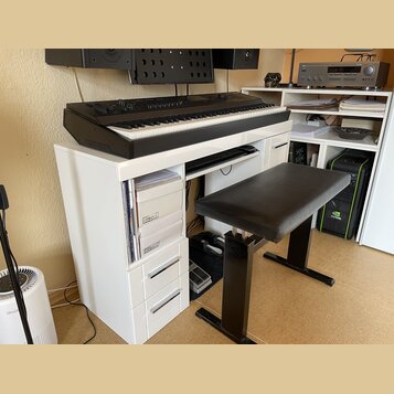 Černý digitální klavír umístěný na bílém stole s přihrádkami. Na levé straně klavíru je nápis MONTREUX. K sestavě patří černá polstrovaná piano stoli…
