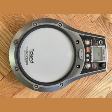 Šedý elektronický tréninkový pad značky Roland s bílou úderovou plochou. Na ovládacím panelu je digitální displej, nápis RHYTHM COACH a modelové ozna…