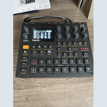 Černý digitální sampler Digitakt II s displejem a ovládacími prvky. Na přední straně je nápis Digitakt II a řada tlačítek, otočných knoflíků a vstupn…