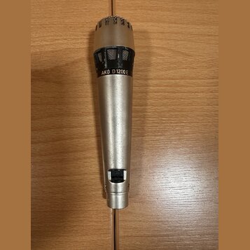 Stříbrný dynamický mikrofon s kovovým tělem a mřížkou. Na horní části je černý nápis AKG D1200E. Povrch těla vykazuje drobné škrábance a známky opotř…