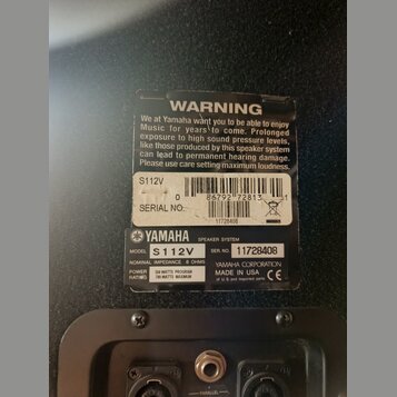 Detailní záběr na zadní stranu černého reproduktoru s dvěma nálepkami. Horní nálepka obsahuje varovný text „WARNING“ a označení „S112V“, čárcový kód…