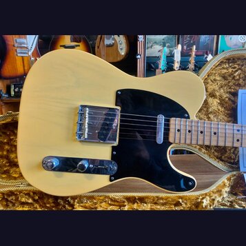 Kytara typu Telecaster v krémové barvě s černým pickguardem a hnědým javorovým hmatníkem. Na kovovém krytu snímače je nečitelný nápis. Nástroj leží v…