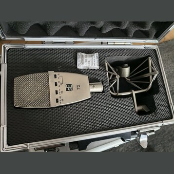 V otevřeném hliníkovém kufru s černou pěnovou výplní je umístěn stříbrný mikrofon a kovový shock mount. Na těle mikrofonu je vidět logo SE a označení…