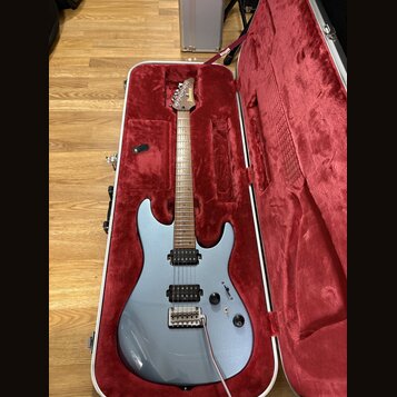 Stříbrná elektrická kytara v otevřeném tvrdém kufru s červeným vnitřkem. Na hlavě hředu je bílý nápis Ibanez. Kytara má dva humbuckery, dva ovládací…