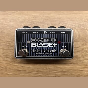 Kytarový pedál v kovovém stříbrném šasi s černým čelem. Na povrchu jsou nápisy SWITCH BLADE+, electro-harmonix a NEW YORK CITY, USA. Jsou zde dva kov…