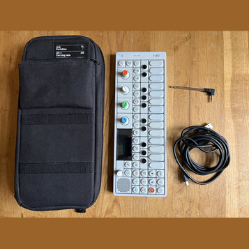 Na dřevěném stole je bílý syntezátor OP-1 s řadou ovládacích knoflíků v různých barvách, displejem a tlačítky. Vedle něj je černý textilní přepravní…