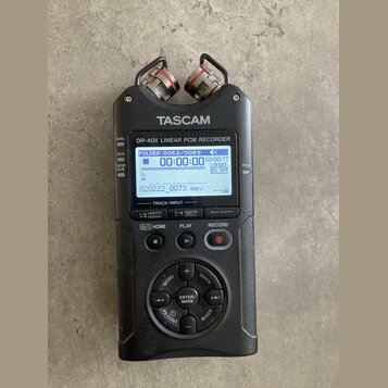 Černý digitální nahrávač s nápisy TASCAM a DR-40X Linear PCM RECORDER. Zařízení má LCD displej s zobrazením času a vlnové formy, horní mikrofony a ov…