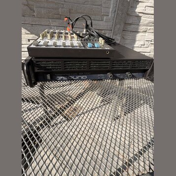 Na černém kovovém mřížkovém povrchu stojí černý rackový zesilovač s nápisem Professional Power Amplifier QSA 1000 a logem Q SOUND. Na zesilovači je u…