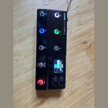 Černý kytarový procesor Line 6 HX STOMP XL s kovovým šasi. Zařízení má osm stoupacích spínačů, z nichž dva jsou podsvíceny modře a zeleně, a jeden če…