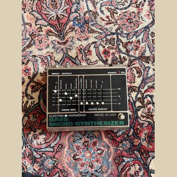 Stříbrný efektový pedál s černým ovládacím panelem. Na spodní části je nápis ELECTRO HARMONIX MADE IN USA BASS MICRO SYNTHESIZER. Panel obsahuje něko…